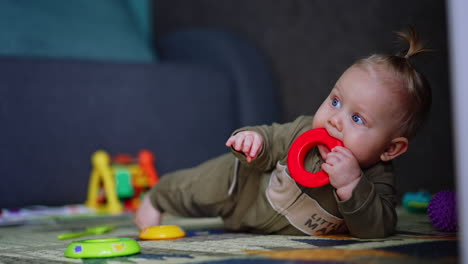 infant child in a romper lies on the floor. cute baby picks a toy and takes it to his mouth. blurred backdrop.