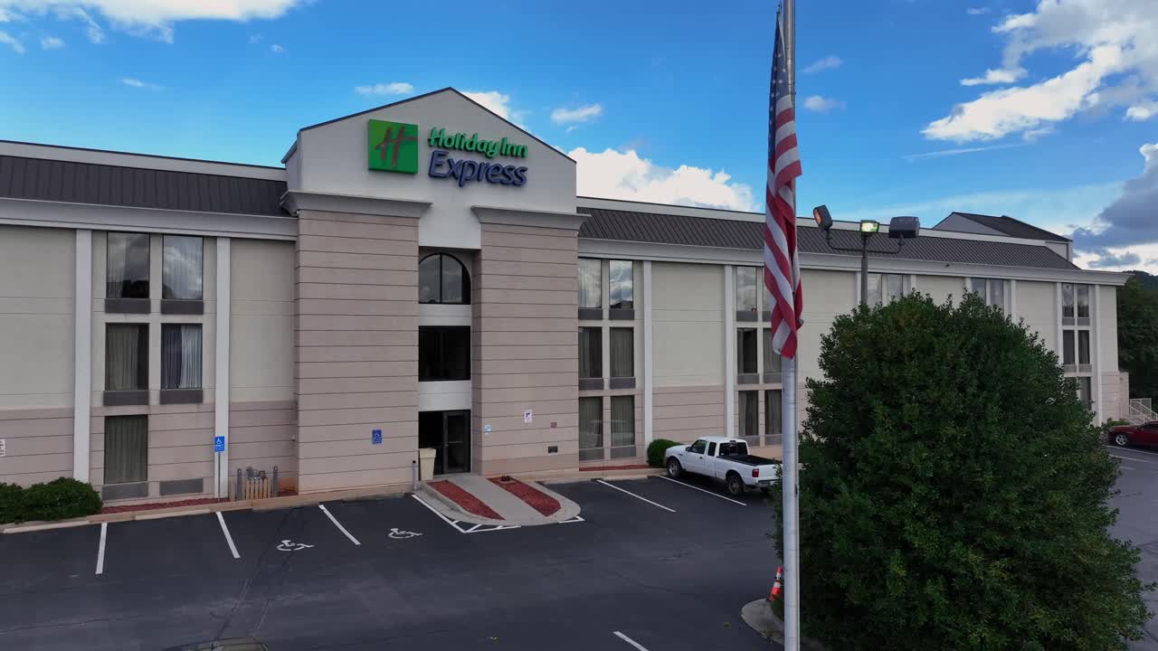 Aerial rising of American holiday inn express hotel in suburb during cloudy day. Green mountains and highway in background. Virginia, USA. Wide shot. American flag in foreground.