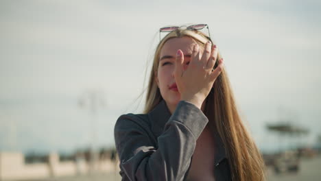 white lady adjusts sunglasses resting on her head with a focused expression, then adjusts her hair, with a blurred background featuring light poles in an outdoor urban setting on a sunny day