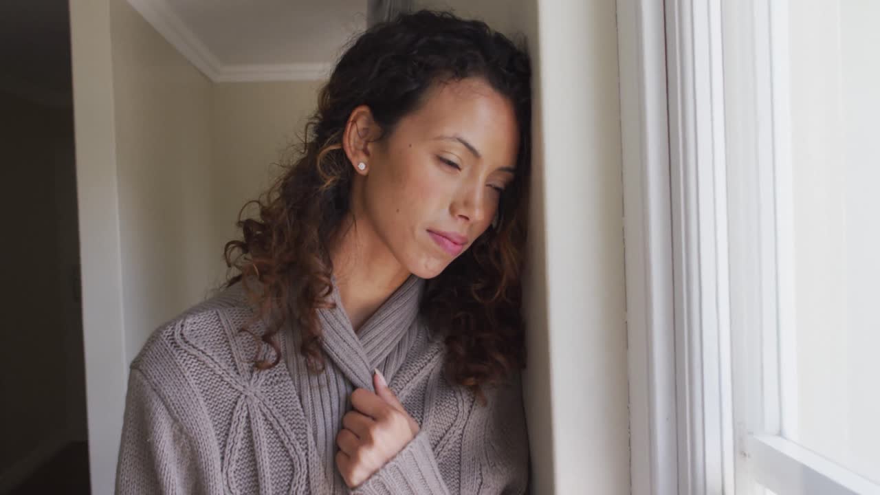 mujer biracial pensativa de pie en la ventana y mirando a la distancia