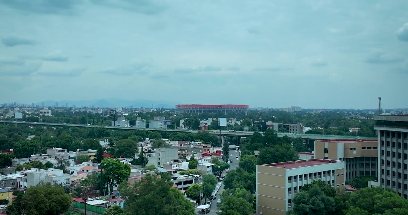 Ampia veduta fissa della zona sud di Città del Messico, con lo Stadio Azteca, infrastrutture stradali sopraelevate e quartieri residenziali sotto un cielo nuvoloso dalle tonalità fredde.