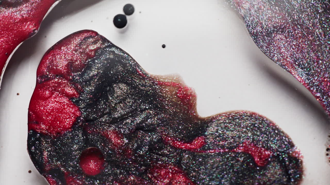 Macro shot of glittery red and black ink droplets moving and merging in a liquid solution as air is blown across them