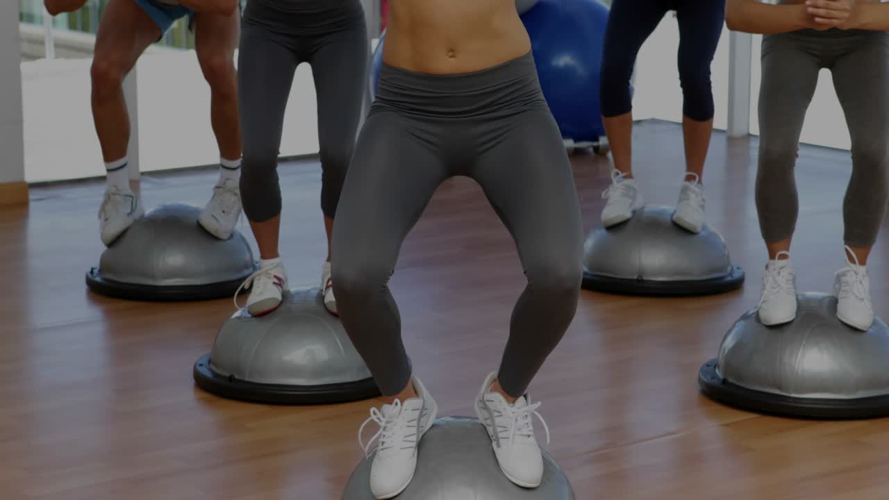Instructor cueing squats, fitness class training balance on BOSUs while overlays sliding over group