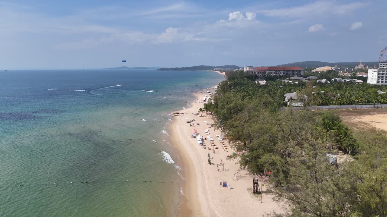 An aerial video shows a pristine beach with gentle waves by a famous resort. Lush trees shade the shore, leading to a tranquil blue lagoon with minimal waves.