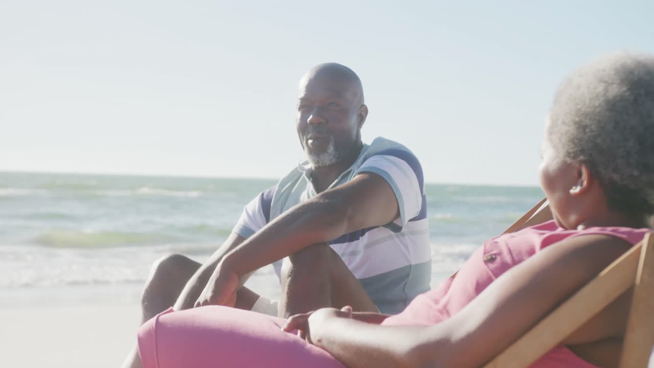 Happy senior african american couple sitting on deck chairs and talking at beach, in slow motion