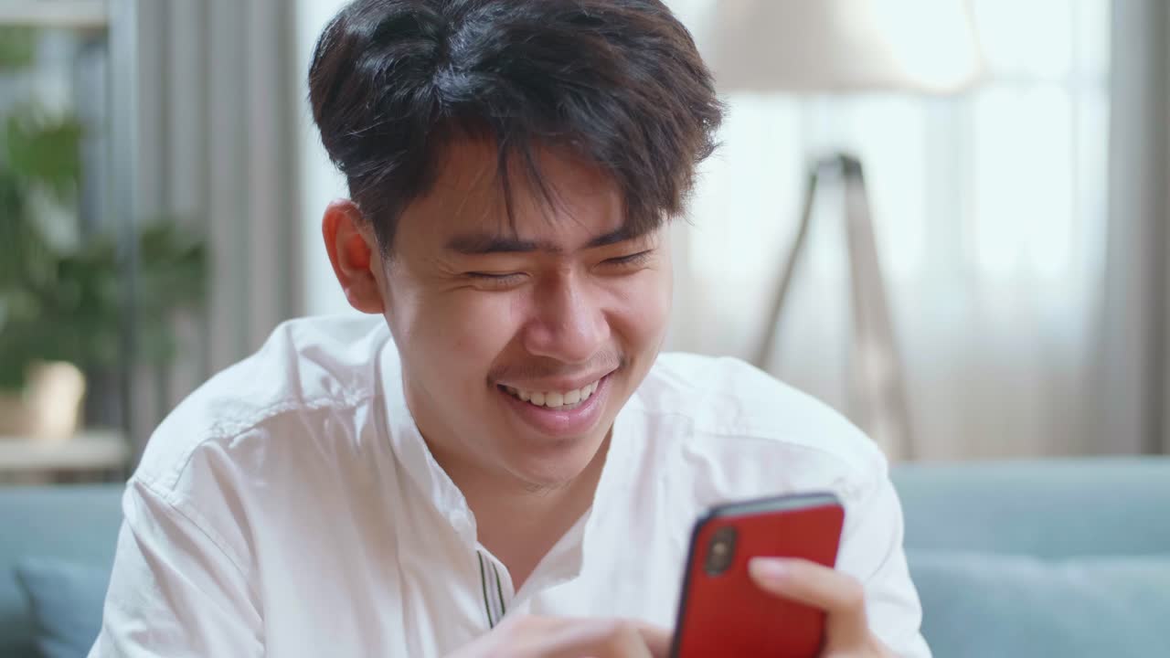 Close Up Of Asian Man Touching Smartphone And Laughing While Sitting On Sofa In The Living Room
