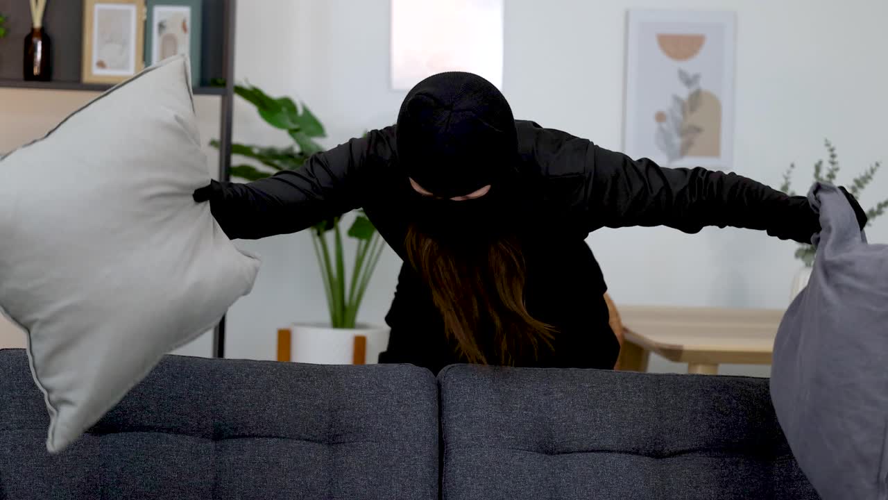 A masked woman in black clothing searches behind sofa cushions in a bright living room