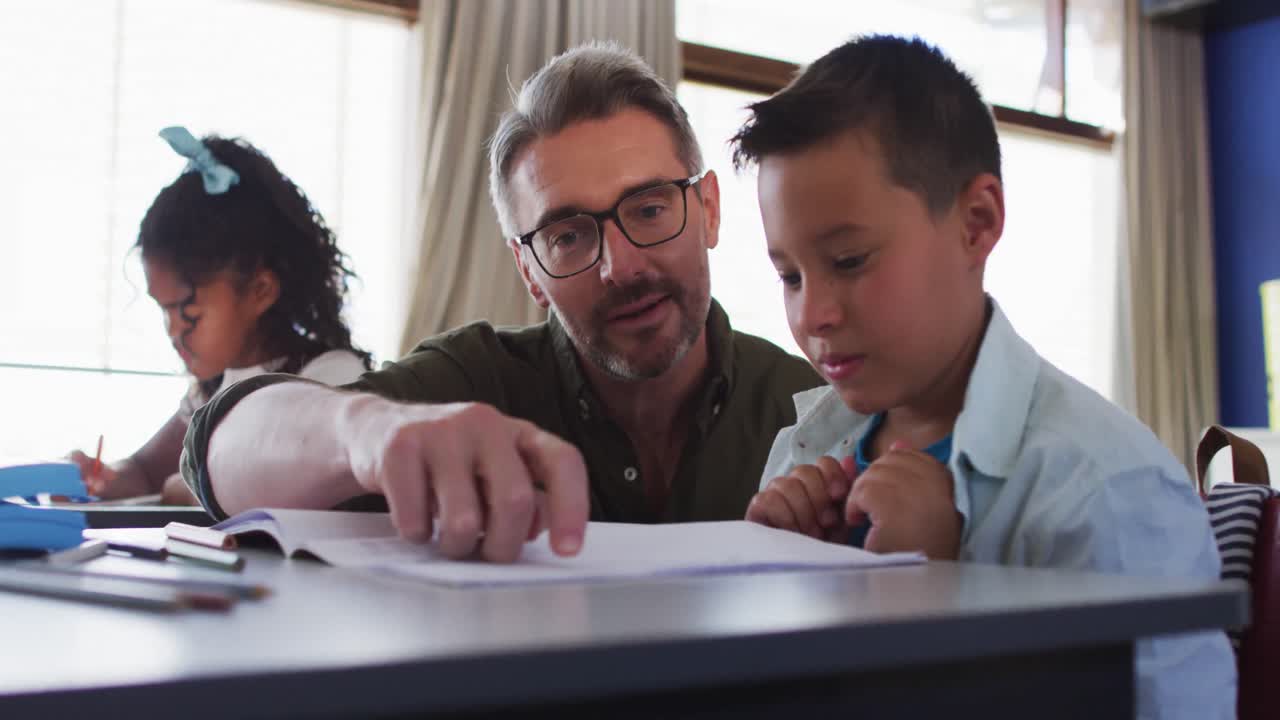 un maestro masculino diverso ayudando a un escolar sentado en el aula a hacer un ejercicio de lectura