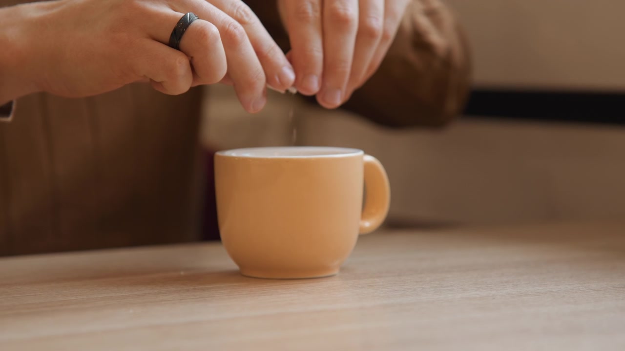 Close up of person adding sugar to orange cup on wooden table. action is performed steadily with hands visible focusing solely on process without additional context or background activity