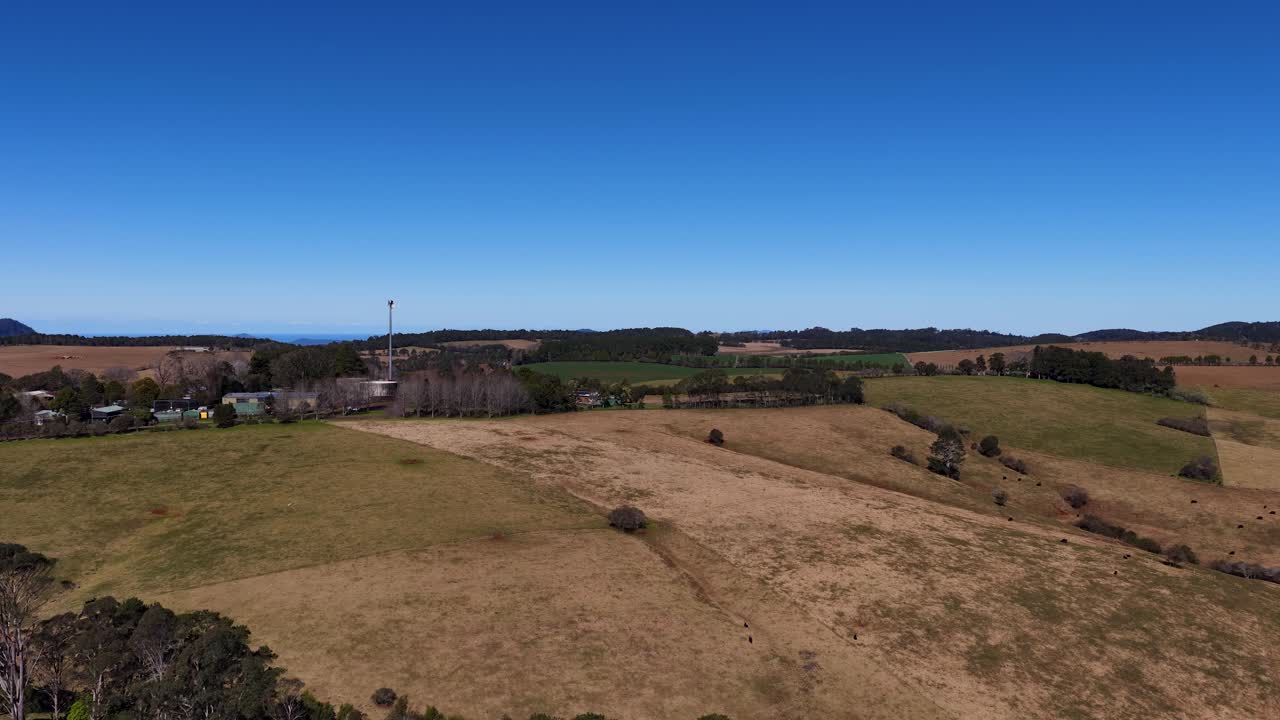 Drone ascends above rural farmland, revealing fields, houses, and distant hills under clear daylight