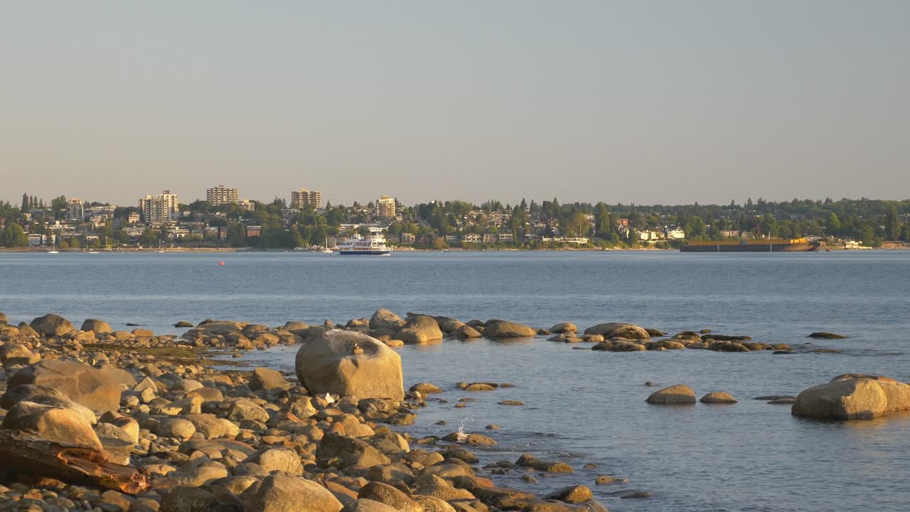 pov desde el paseo marítimo con rocas en la orilla y un ferry navegando en la distancia durante la puesta de sol en vancouver, canadá