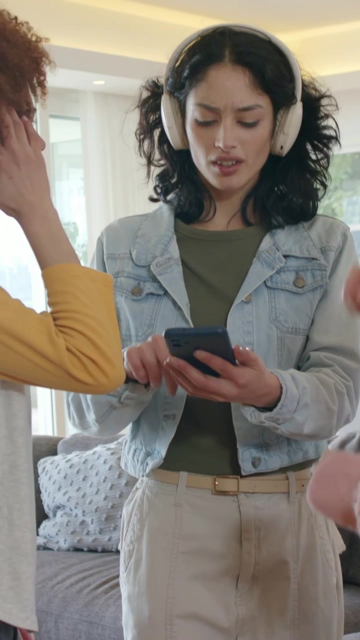 Woman in denim jacket scrolling smartphone after alert in living room, friend in pink knit pointing