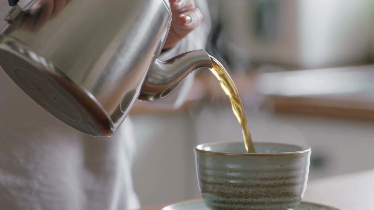 Mani con maglia a maniche lunghe che versano, inclinando un bollitore d'acciaio e riempiendo una tazza sul bancone della cucina, con vapore che sale.