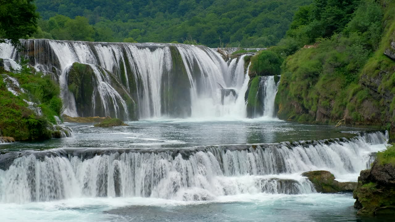 Kayalardan aşağıya doğru akan şelaleler, akan suyla doğal bir sahne oluşturuyor. Arka planda yeşil ağaçlar var. Görüntüde birden fazla seviyede şelale görülüyor.