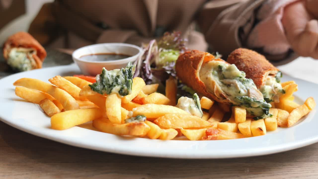 Primo piano di una mano che prende degli involtini fritti con spinaci e formaggio serviti con patatine fritte