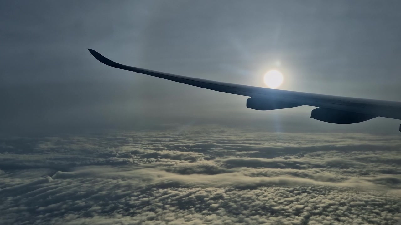 Utsikt fra vindusplass på flyvinge som flyr høyt over et tykt lag med hvite skyer mot en lysende sol som skaper linseblending på himmelen