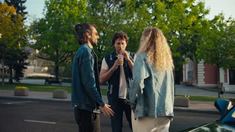 A-happy-company-of-young-friends-in-denim-jackets-are-standing-near-their-gray-cabriolet-and-drinking-yellow-cocktails-on-a