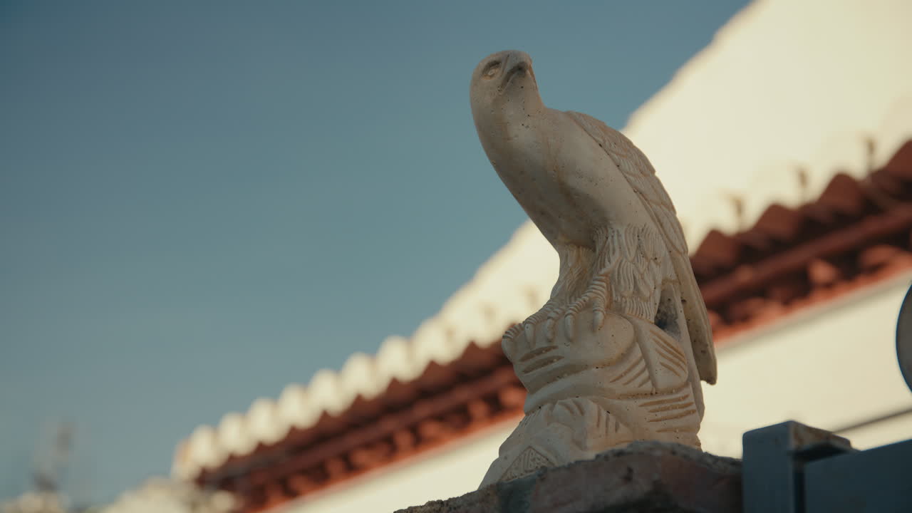 Close-up of carved eagle statue perched on wall, lit by warm sunlight with blue sky and Spanish-style building in the background