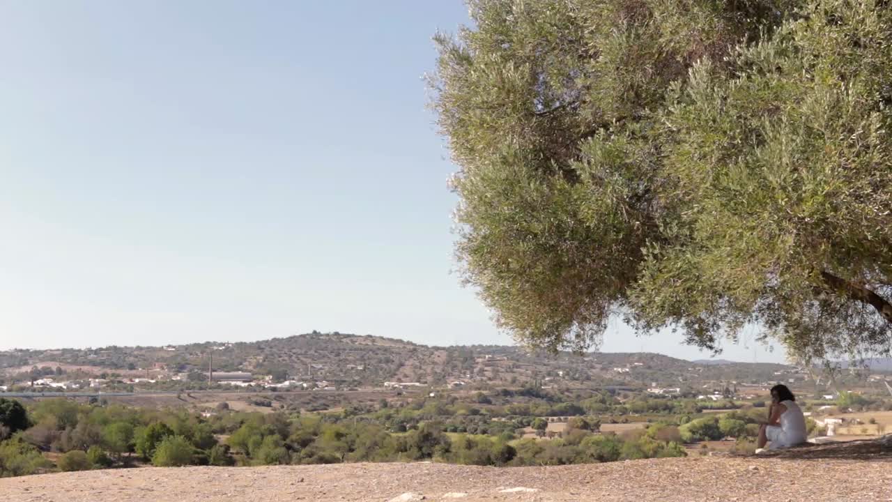 Donna rilassata vestita di bianco seduta sotto un antico ulivo con vista su una catena montuosa verde.