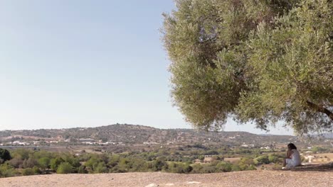 relaxing woman dressed in white sat under ancient olive tree overlooking green mountain range.