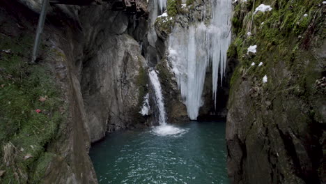 Drone-flying-towards-small-waterfall-and-blue-water-lake-between-cliffs-with-moss-and-icicles-in-Austria-at-the-Sigmund-Thun-Klamm