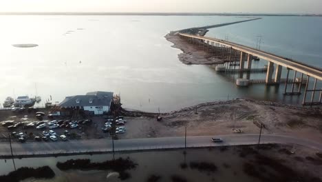 Sobrevuelo-Aéreo-De-Un-Restaurante-Romántico-Con-Vistas-A-La-Puesta-De-Sol-En-Laguna-Madre-Cerca-Del-Puente-En-La-Autopista-Jfk-Memorial-En-La-Isla-Del-Padre-Norte-Cerca-De-Corpus-Christi,-Texas