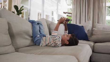 niño biracial feliz acostado en el sofá y usando la tableta en casa, cámara lenta