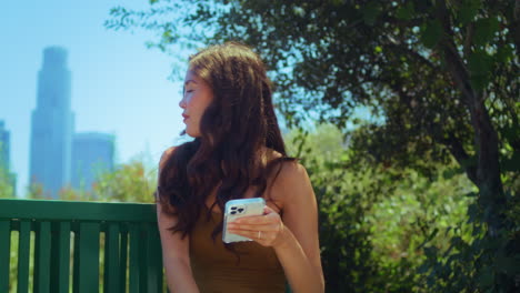 Asian-woman-looking-back-on-blurred-cityscape-holding-smartphone-in-park-closeup