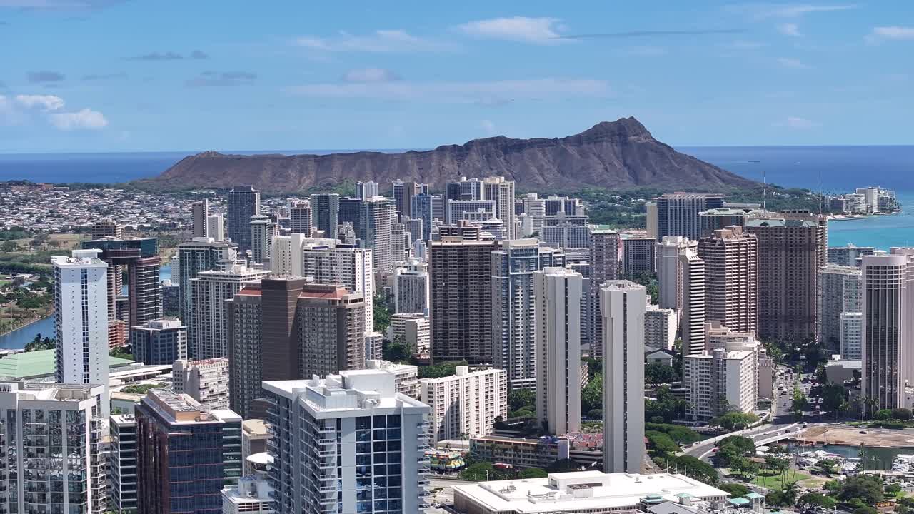Aerial drone footage of Honolulu skyline on Oahu, Hawaii, with Diamond Head volcanic crater in background, showing high-rise hotels, tropical coastline, turquoise waters, and scenic island landscapes