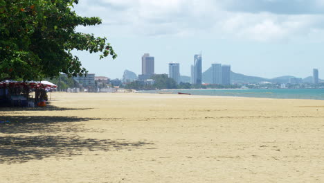 pattaya beachfront with big umbrellas on the left then the beautiful clean sand, shoreline with boats and waves rolling, in the horizon is the city revealing hotels, condos, and the mountains