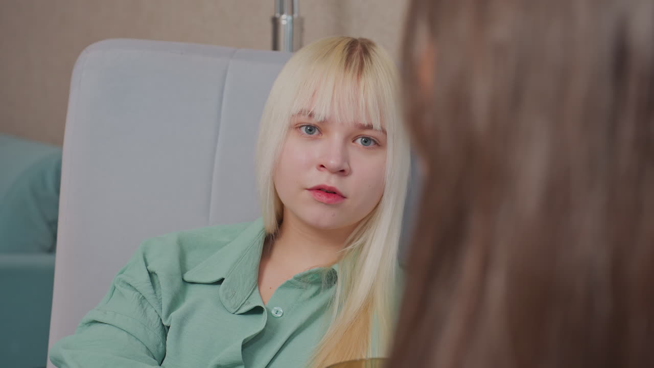 Blonde girl in green shirt reclines against cushioned headboard while holding cup and engaging in casual conversation with unseen friend, indoor environment and soft background