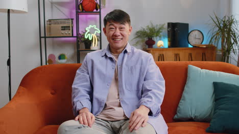 asian man smiling friendly at camera, waving hands gesturing hello, hi, greeting at home on sofa