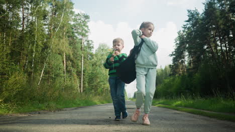Children walking calmly along tarred road surrounded by green forest as girl adjusts bag strap and boy holds wildflower, enjoying quiet stroll under soft daylight and clear blue sky