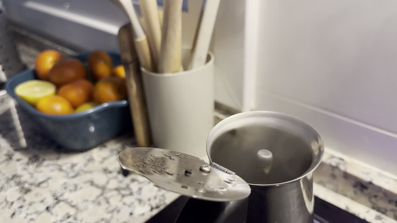 Moka pot steaming on stove with fruit bowl and utensils in cozy kitchen setting