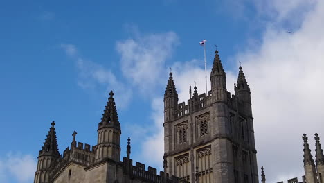 Super-Slow-Motion-Shot-of-Seagull-Flying-in-and-out-of-Frame-with-Tower-of-Bath-Abbey-in-Somerset,-England-in-the-Background-on-Sunny-Summer’s-Day-with-Saint-George’s-Cross-Flapping-in-the-Wind