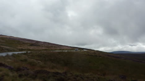 Forwards-fly-along-road-and-tracking-silver-car-on-cloudy-day.-Narrow-curvy-route-through-highlands.-Ireland