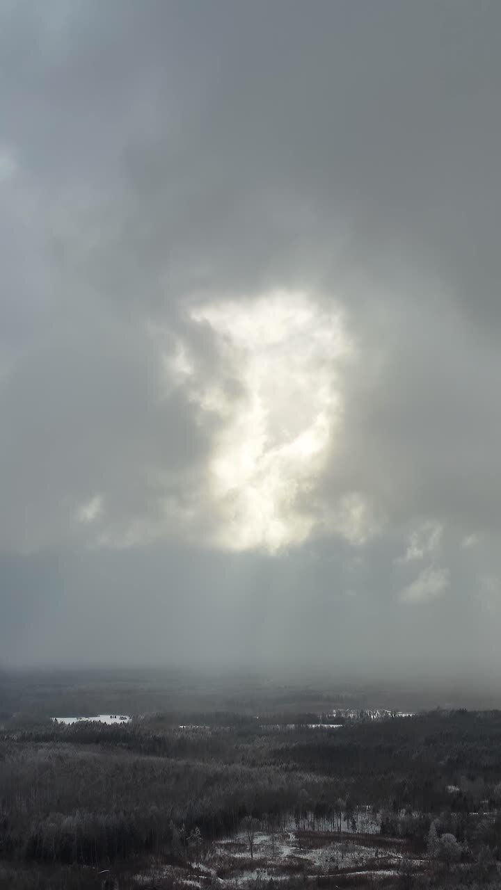 Sun rays breaking through thick clouds in winter. Vertical aerial view over the countryside landscape. Snow storm approaching. Dramatic sky over the forest.