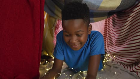 happy african american boy using tablet and smiling lying in blanket camp