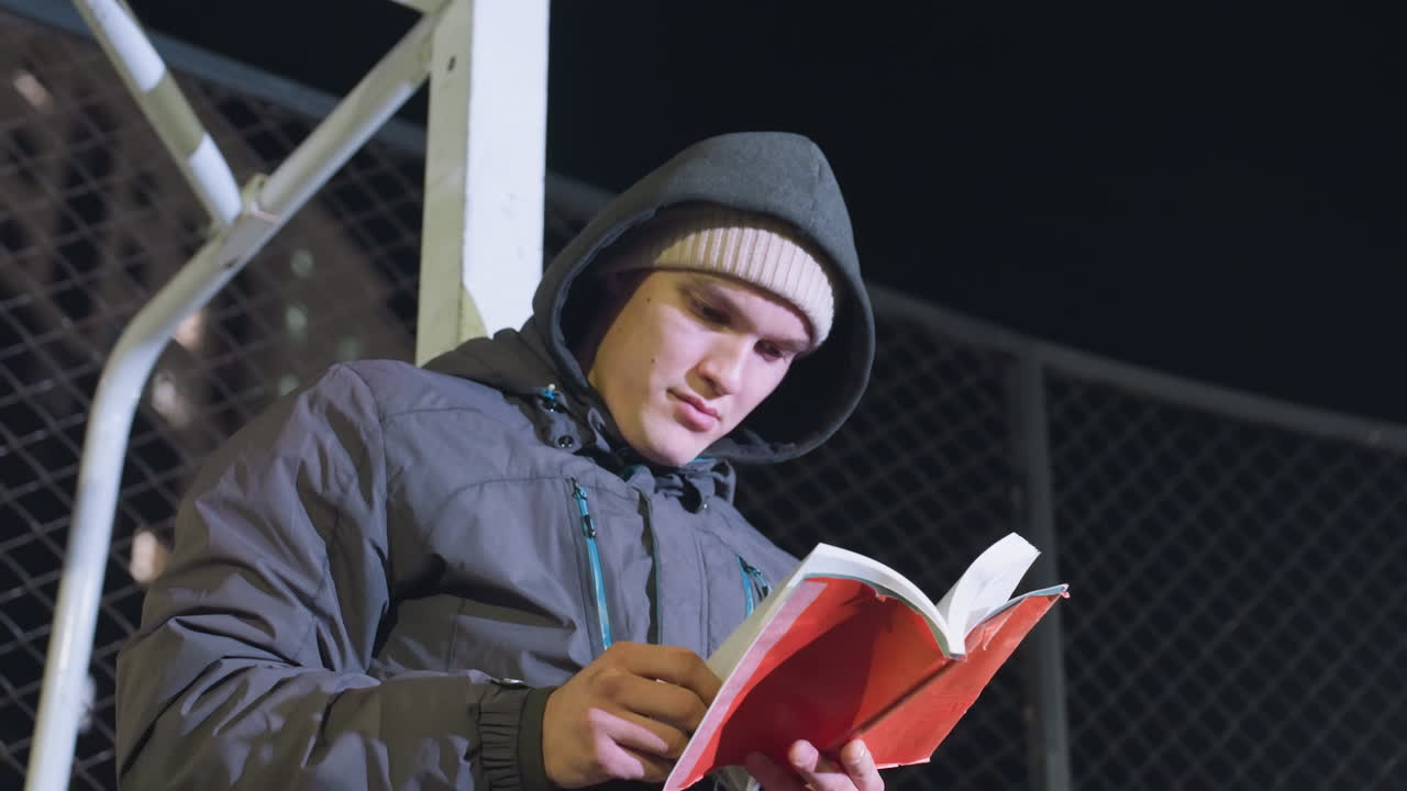 Young man in hoodie and beanie leaning against metallic goalpost, focused reading book outdoors at night with mesh fence background