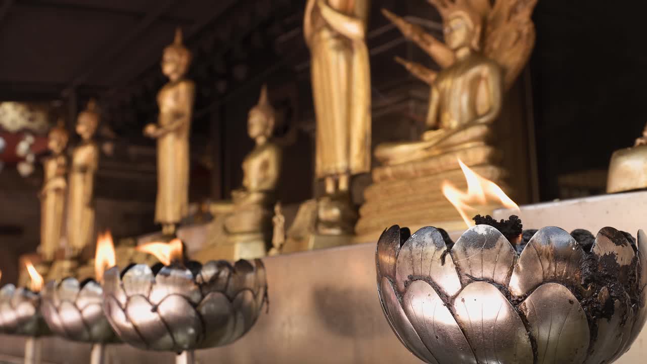 Close-up shot of candles with moving flame in lotus-shapes candle holders with row of golden Buddha statues on the background in a Buddhist temple