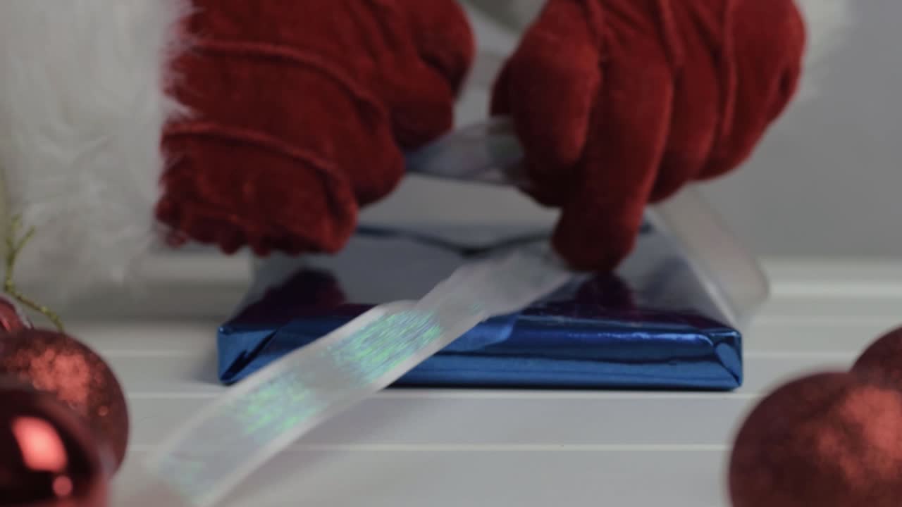 Hand wearing Santa gloves wrapping Christmas gift with ribbon and blue paper close up shot