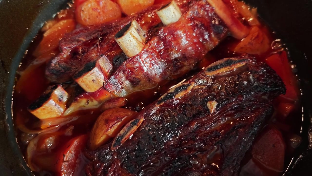 Vista dall'alto di costine di manzo Asado a cottura lenta in brodo ricco con patate, patate dolci e cipolla che bollono in una pentola nera. Stufato di carne gourmet rustico, primo piano di comfort food.