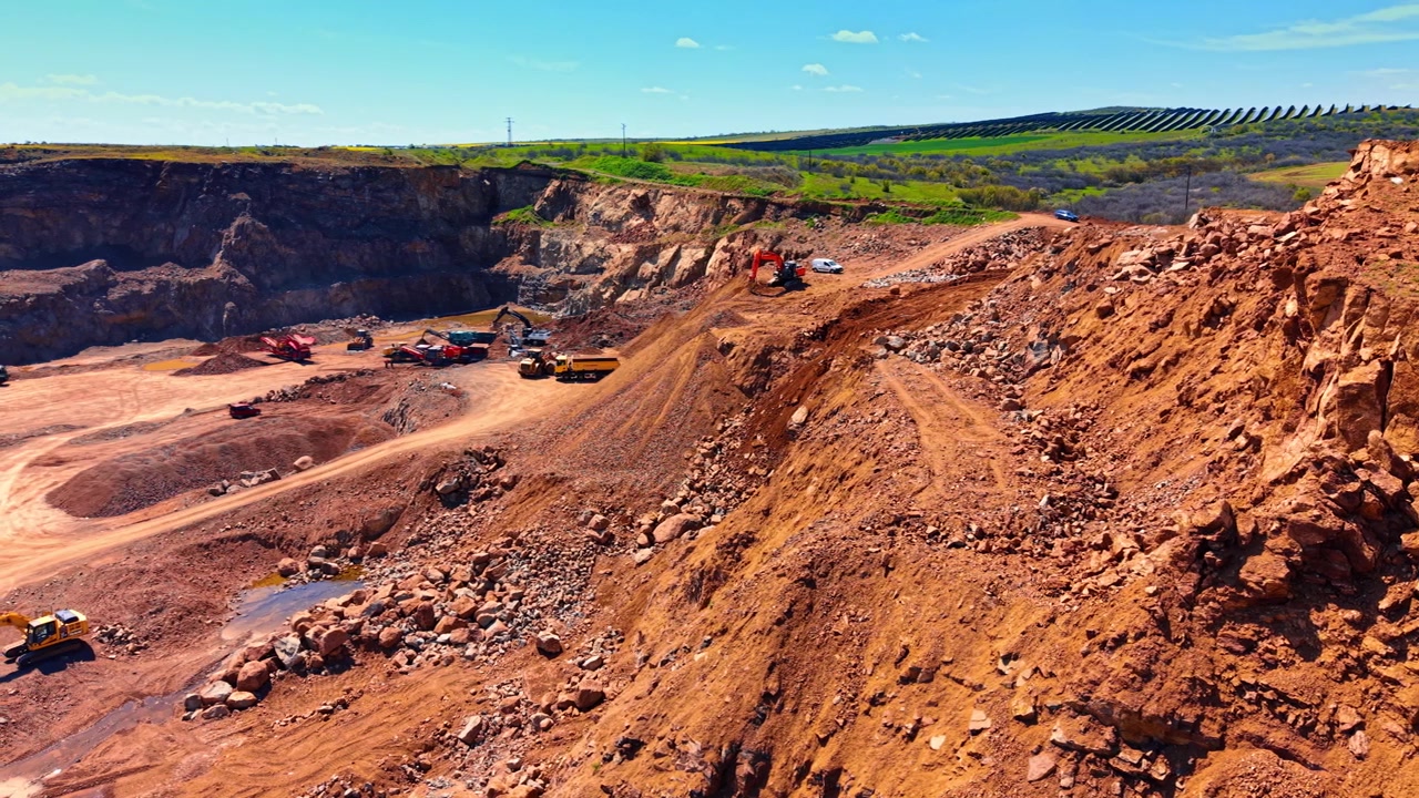 Open stone quarry with terraced rocky slopes and solar farm horizon. High angle perspective shows red earth slopes and machinery with green fields and solar panels under blue sky.