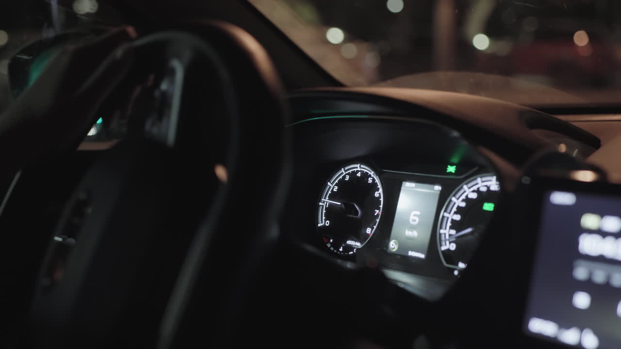 Close up of driver turning steering wheel inside moving car at night with illuminated dashboard, digital speedometer, and blurred street lights visible through windshield