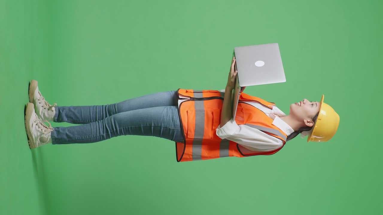 Full Body Side View Of Asian Female Engineer With Safety Helmet Working On A Laptop And Looking Around While Standing In The Green Screen Background Studio