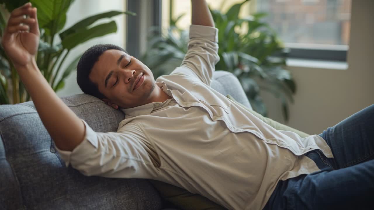 Uomo che si stira al risveglio, allungando gradualmente le braccia su un divano grigio, indossando una camicia chiara e jeans