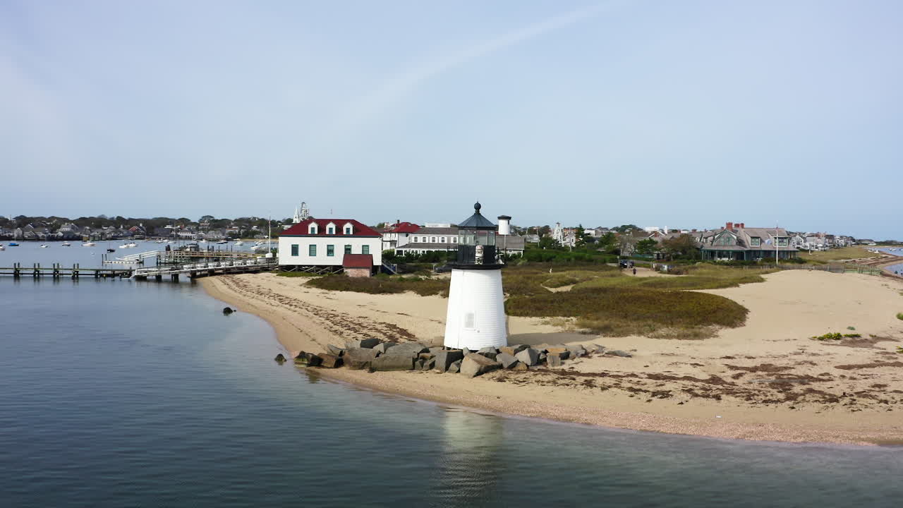 Drone che orbita a sinistra oltre il Faro di Brant Point a Nantucket, Massachusetts, rivelando case costiere, spiaggia sabbiosa e acque tranquille del porto in una ripresa aerea costiera cinematografica.