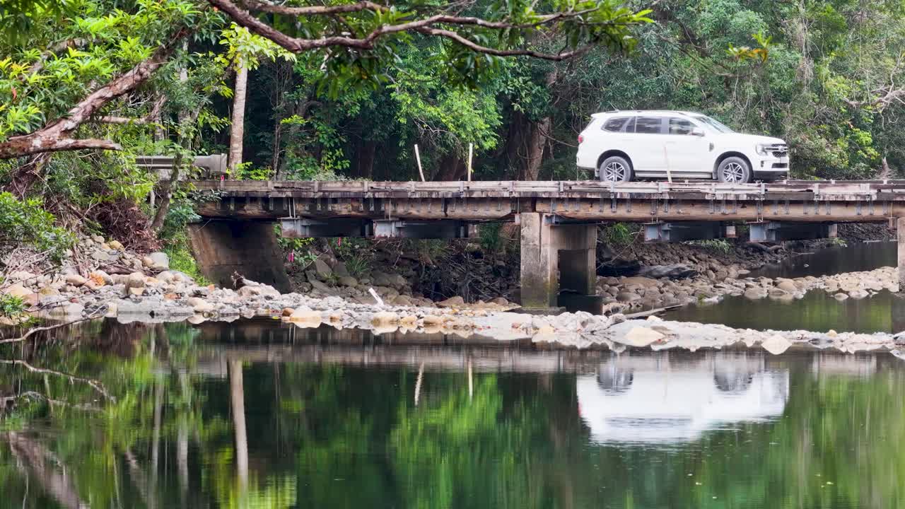 A white SUV crosses a rustic bridge over a calm creek in a dense rainforest, reflecting serene natural beauty