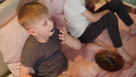 aerial view of little children sitting on pink bed enjoying fresh fruits from brown glass plate cozy indoor setting with boy reaching for fruit while girl sits nearby