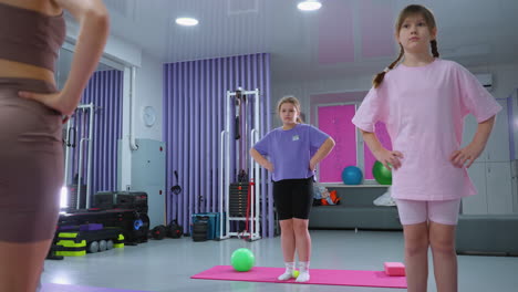 vista parcial de un instructor de fitness dirigiendo a estudiantes jóvenes en ejercicios de construcción de resistencia, manos sobre su cintura, en un gimnasio con esteras y equipos rosados en el fondo